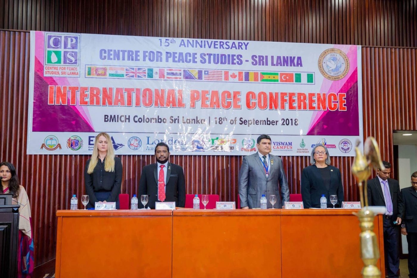 The conference panel observing a moment of silence at the head table, beneath the 15th-anniversary banner